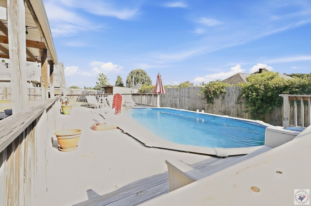 Backyard swimming pool beside a raised wooden deck and covered porch, enclosed by a wooden privacy fence.