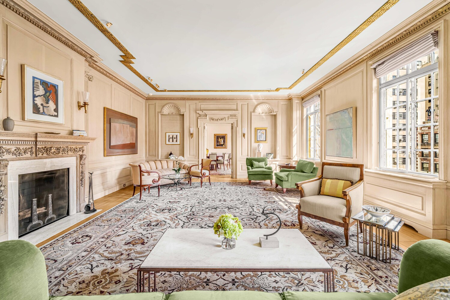 Living room with ornate cream paneling, a carved fireplace, green armchairs, and large city-facing windows.