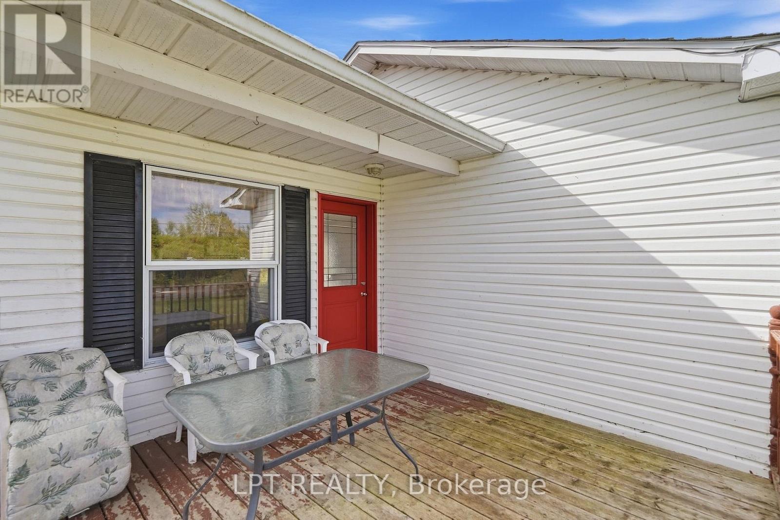 Covered deck with weathered wood flooring, outdoor chairs, and a glass-top table beside a red door.