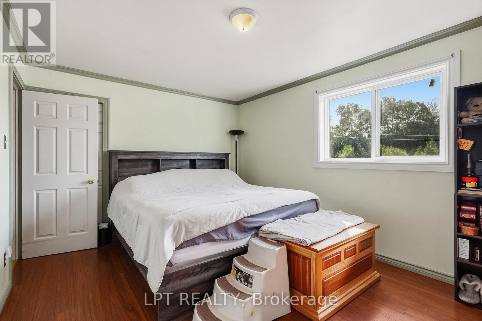 Bedroom with a large bed, hardwood floors, and a wide window overlooking trees.