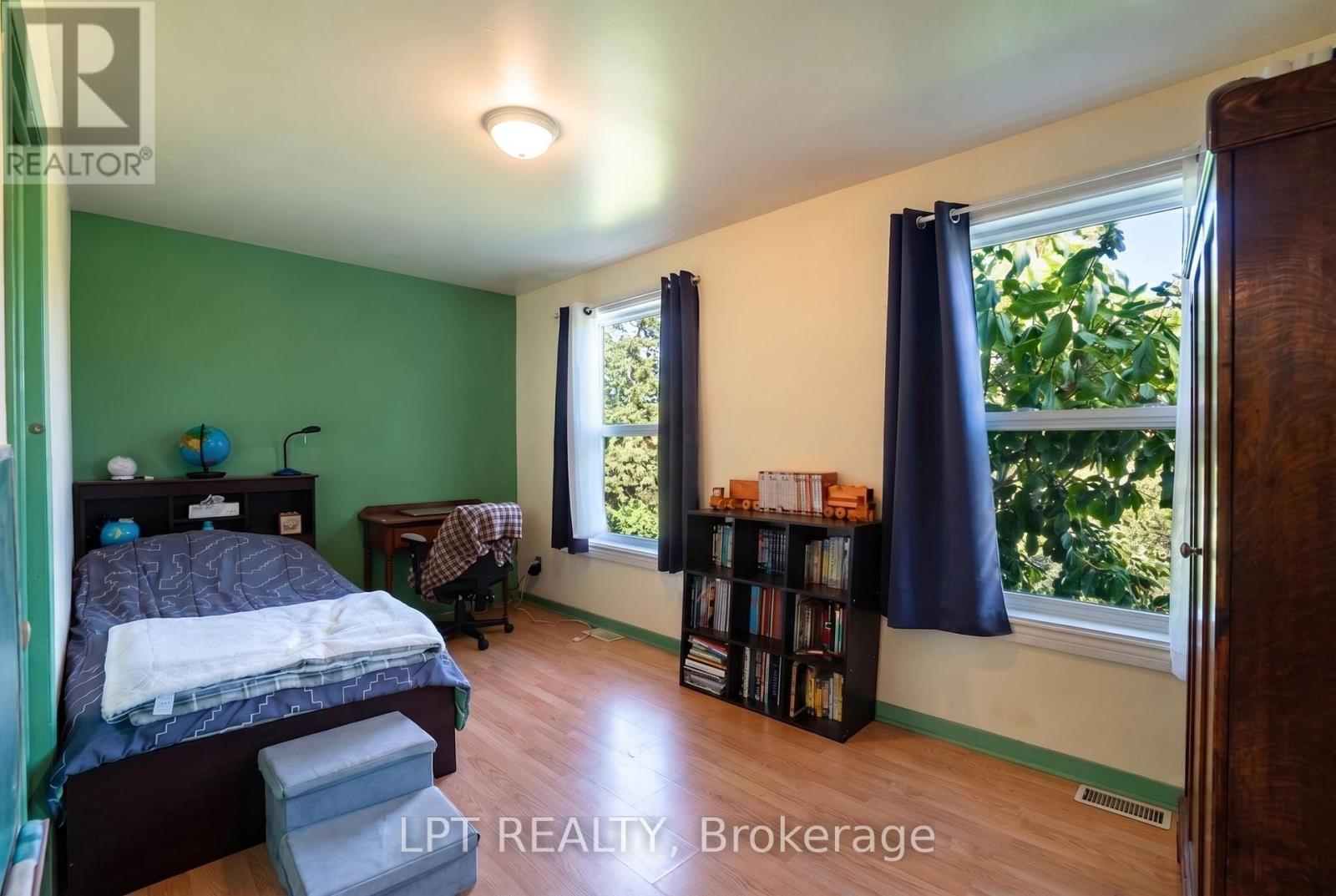 Bedroom with a green accent wall, bed, desk, and tall windows overlooking leafy trees.