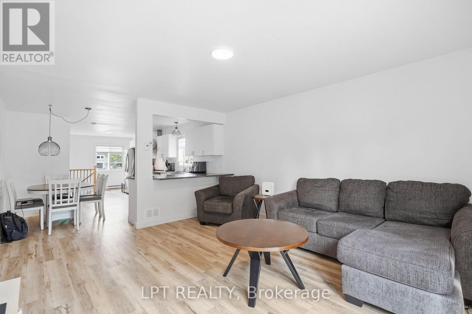 Open-plan living room with a gray sectional, round coffee table, dining area, and white kitchen.