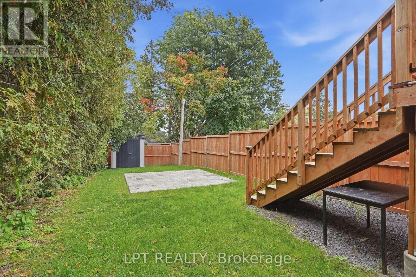 Fenced backyard with a paved patio, dark storage shed, and wooden stairs to a deck.