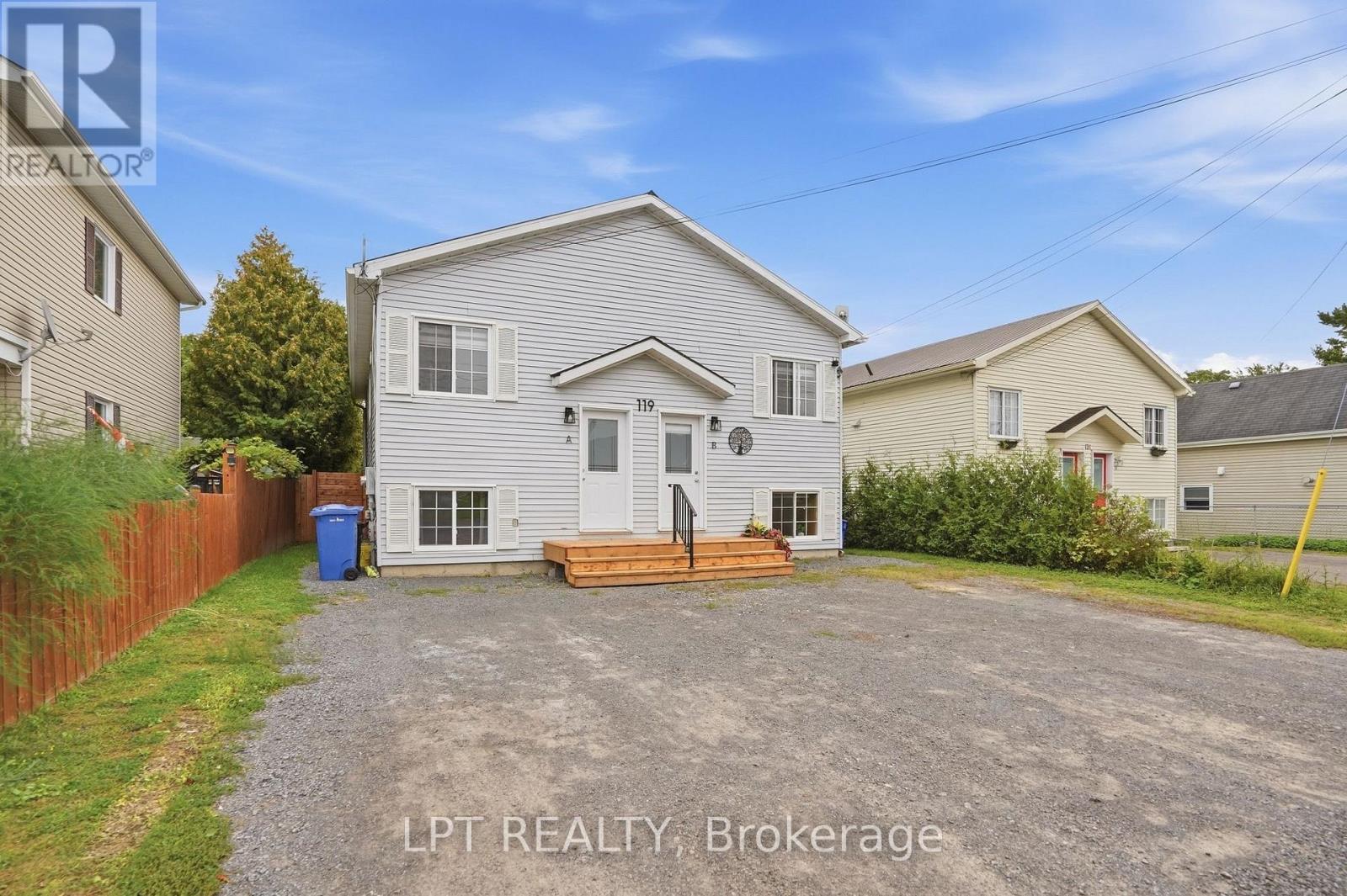 White-sided two-unit house with separate entrances and wooden steps behind a gravel driveway.