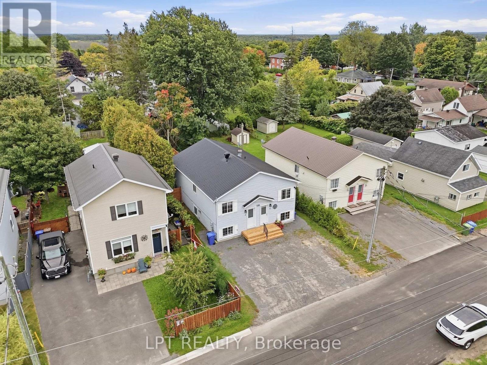 Aerial view of a white house with a front deck, gravel driveway, and tree-lined neighborhood.