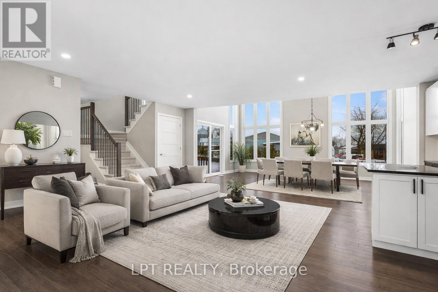 Staged open-concept living and dining room with pale sofas, a staircase, and tall windows.