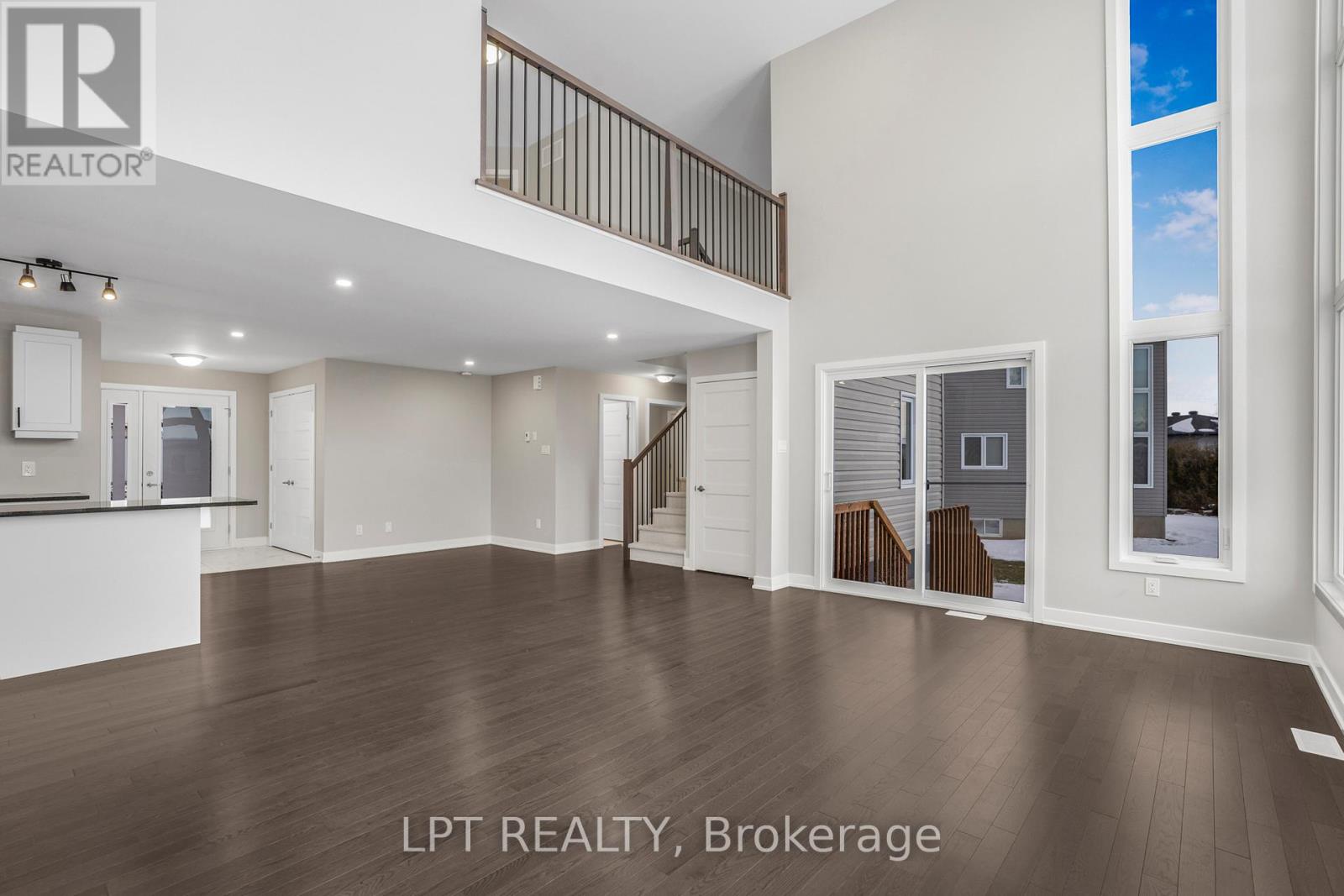 Empty open-concept living area with dark hardwood floors, a staircase, upper-level railing, and tall windows.