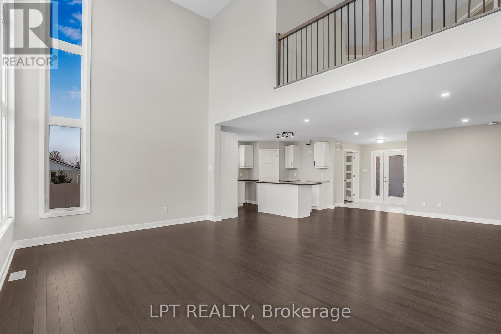 Open living room with dark hardwood floors, soaring ceiling, tall windows, and an adjoining kitchen.