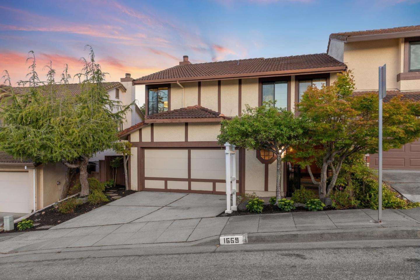 Two-story beige home with brown trim, tiled roof, and landscaped front yard at sunset.