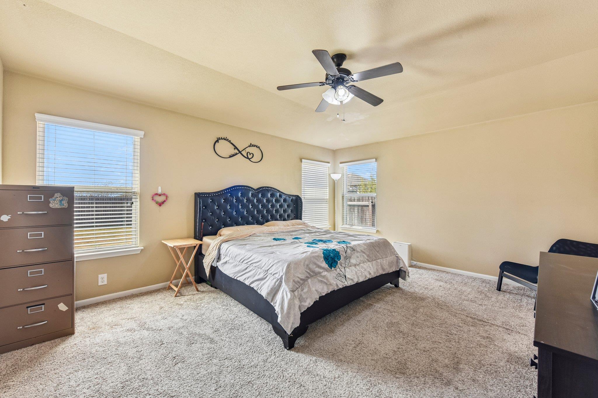 Bedroom with a dark tufted headboard, floral bedding, and three windows with blinds.