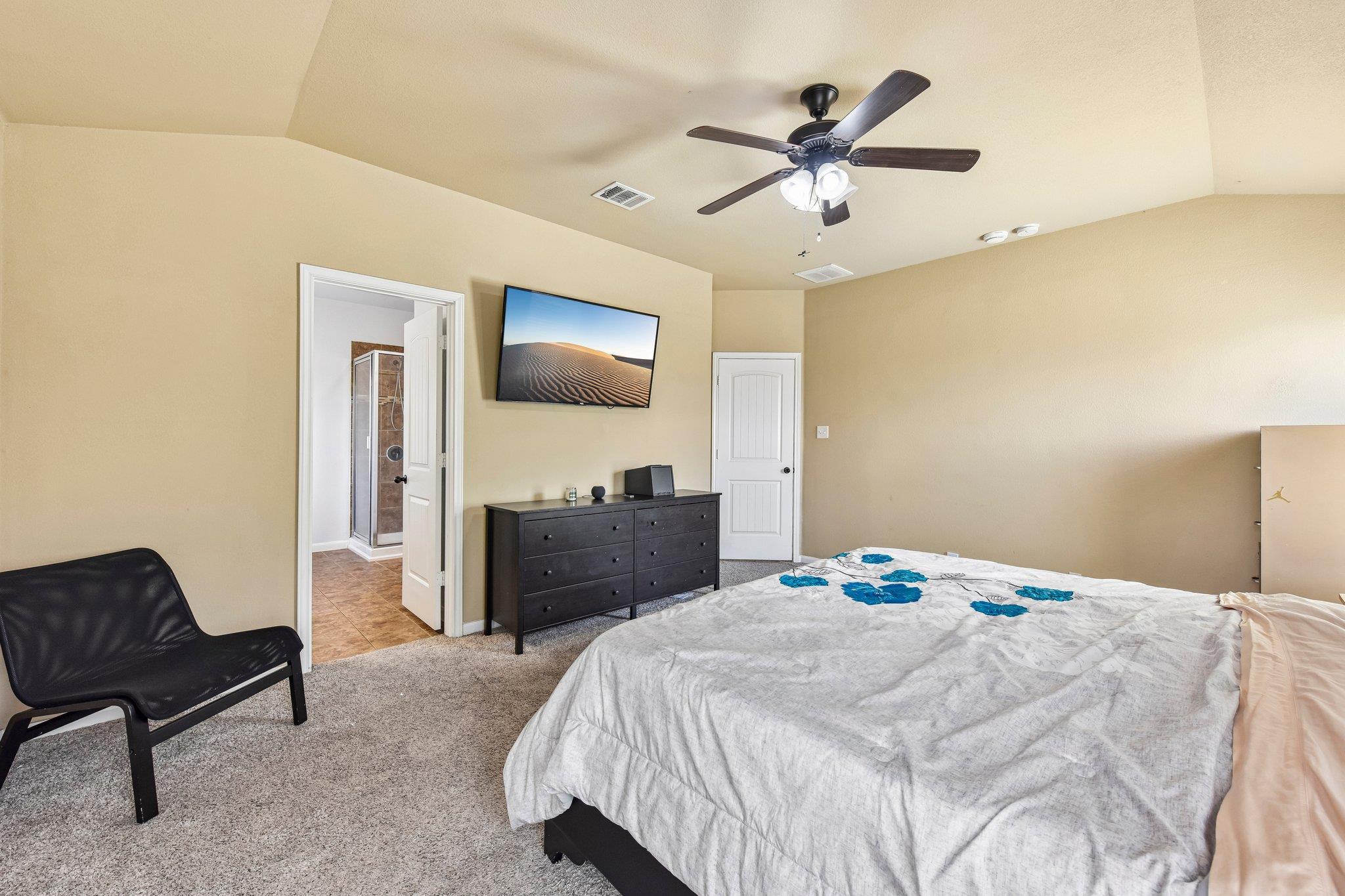 Bedroom with a blue-patterned bedspread, dark dresser, wall-mounted TV, and doorway to a bathroom.