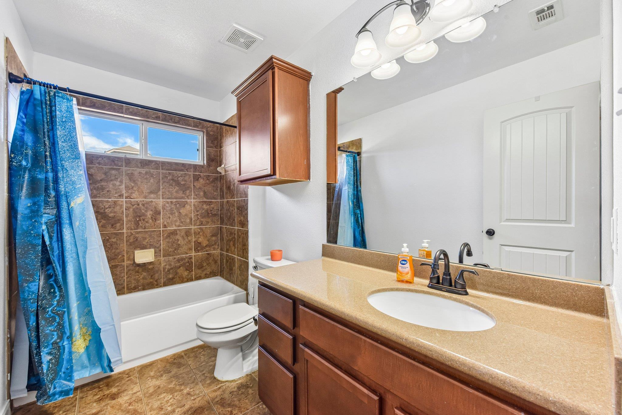 Bathroom with brown tiled tub and shower, blue curtain, and wood vanity with tan counter.
