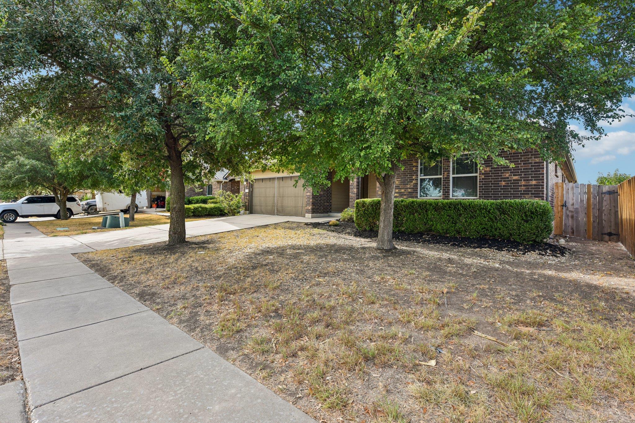 Brick home with a two-car garage behind mature trees, beside a dry front lawn and sidewalk.