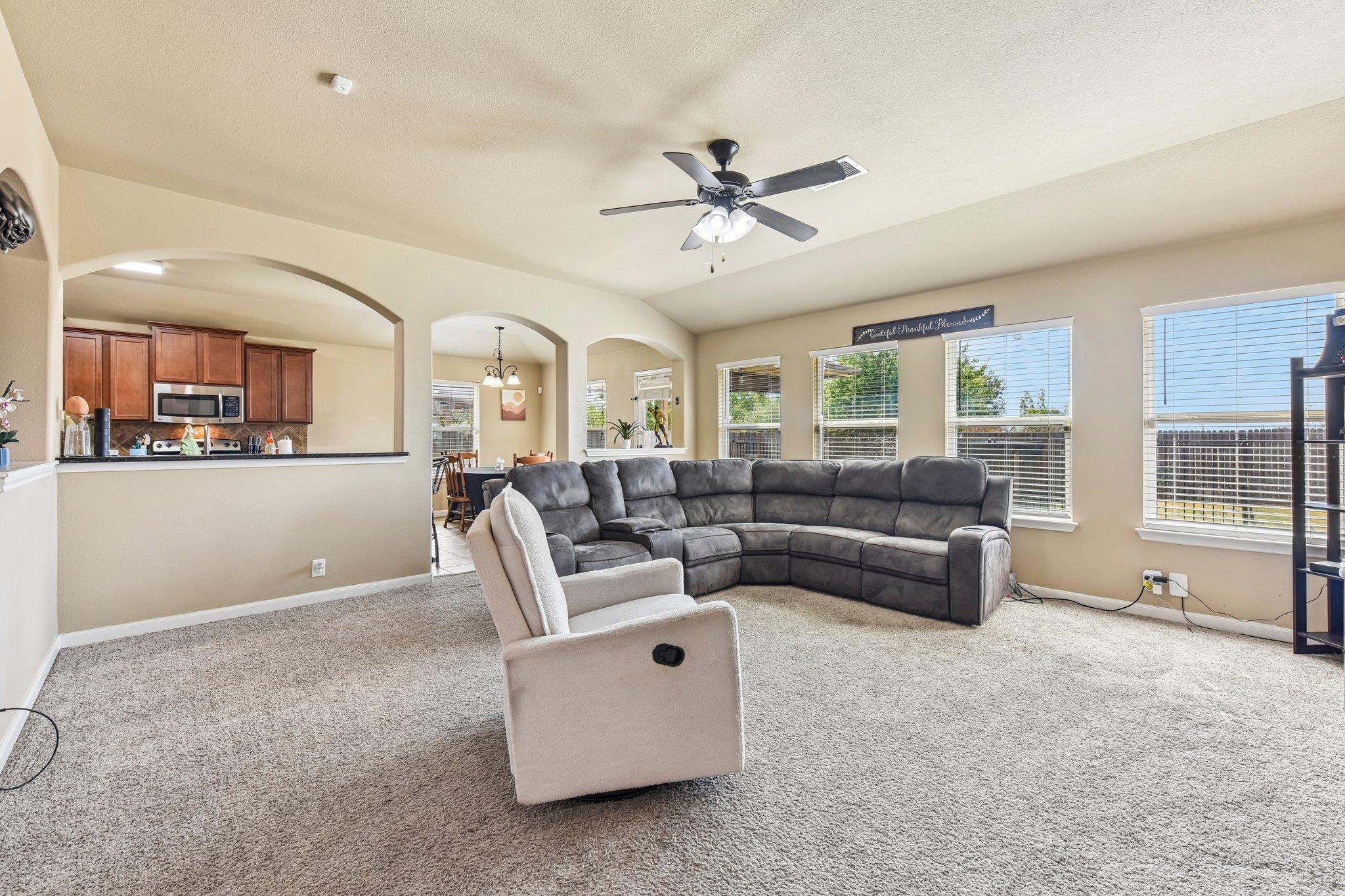 Living room with a gray sectional, beige recliner, carpet, and arched openings to the kitchen and dining area.