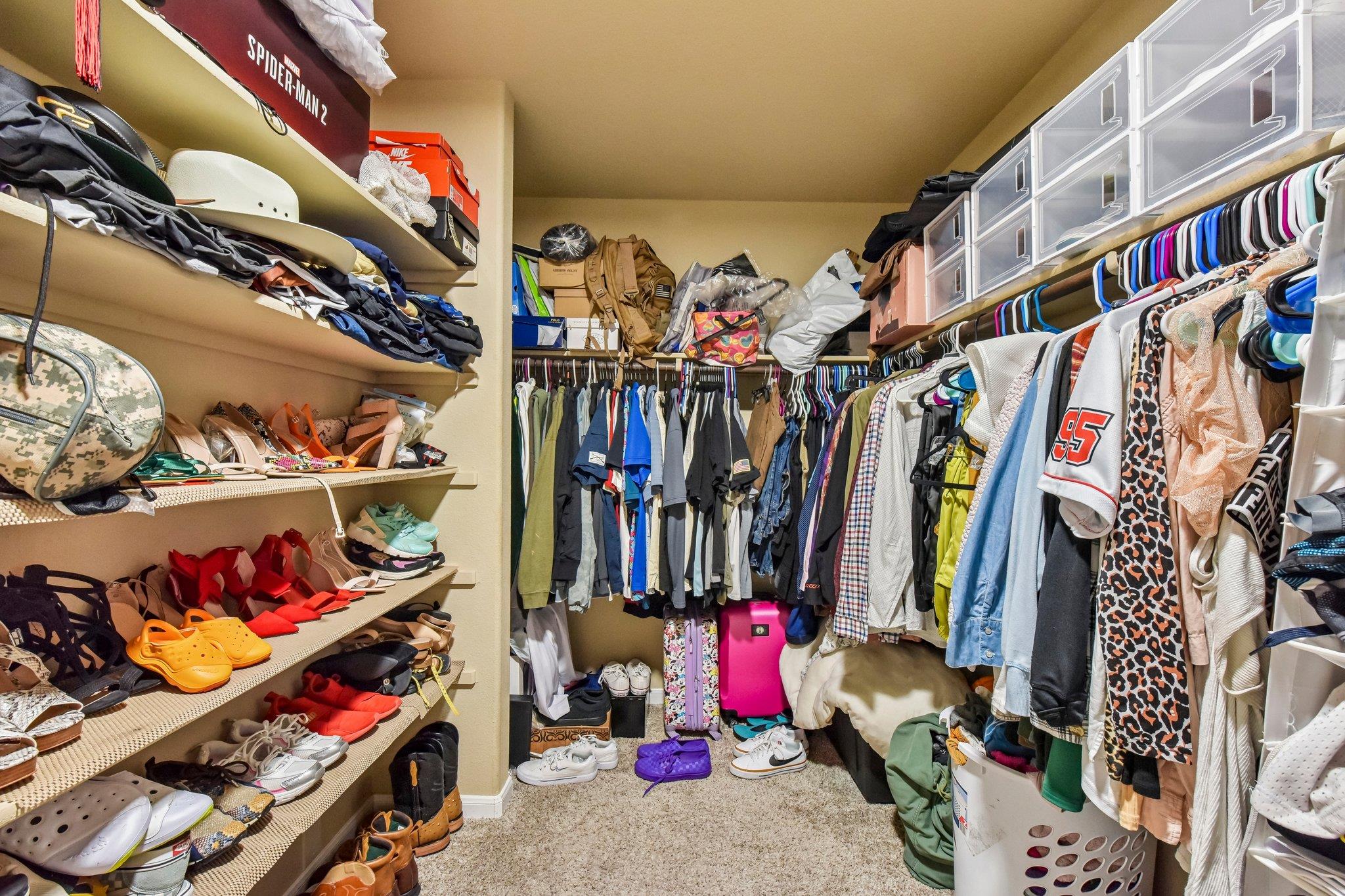 Walk-in closet with extensive shoe shelving, crowded clothing racks, clear drawer organizers, and colorful luggage.