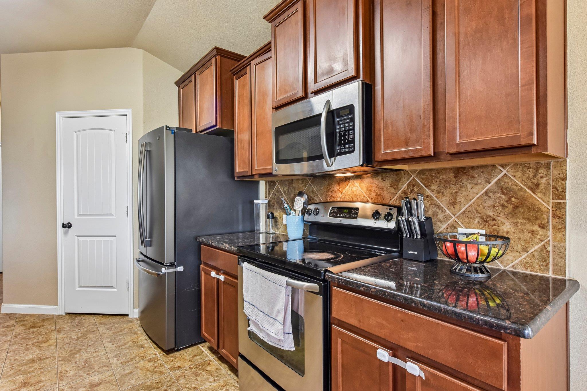 Kitchen with reddish wood cabinets, dark counters, stainless steel appliances, and a tiled backsplash.