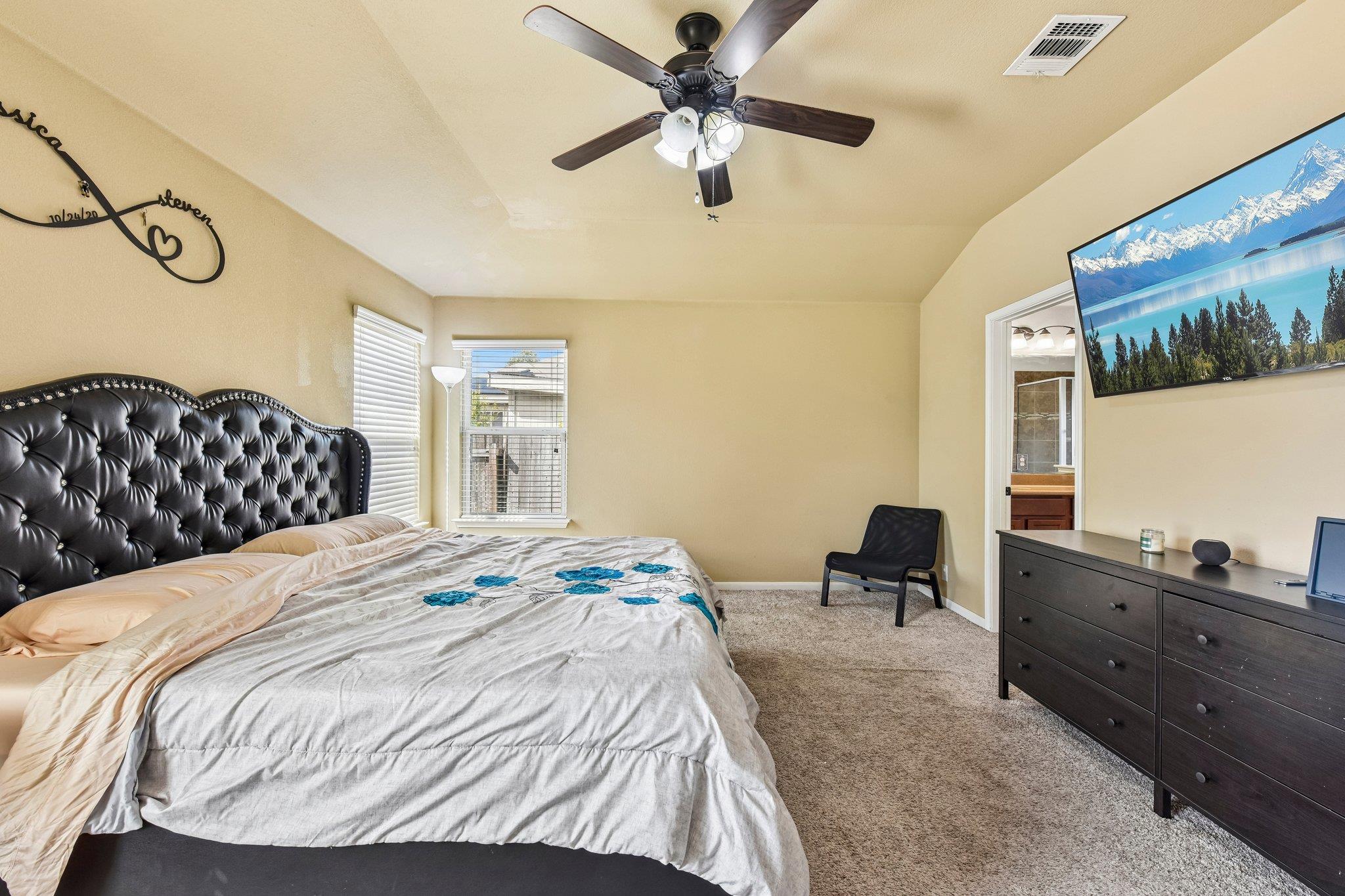 Bedroom with a black tufted headboard, ceiling fan, and doorway to an ensuite bathroom.