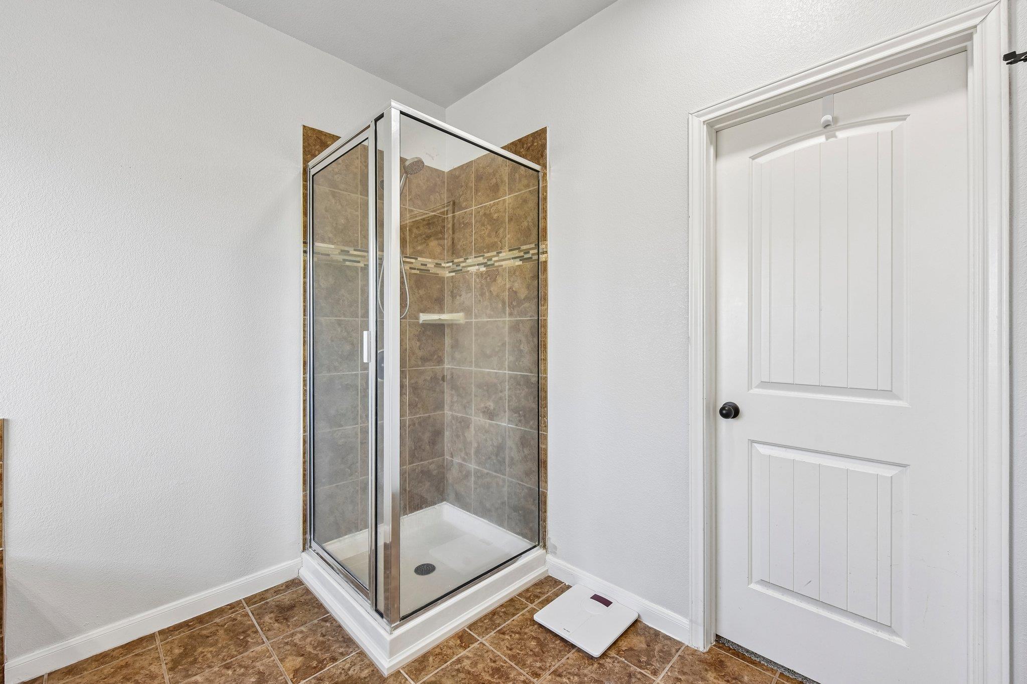 Bathroom corner shower with glass enclosure, brown tile, and a scale beside the door.