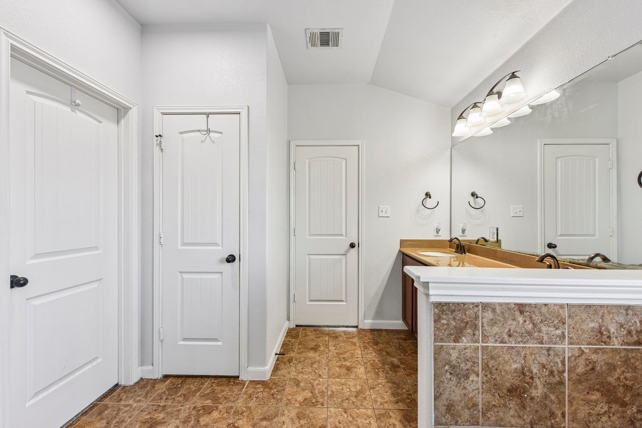Bathroom with a double vanity, large mirror, tan tile floor, and several white paneled doors.
