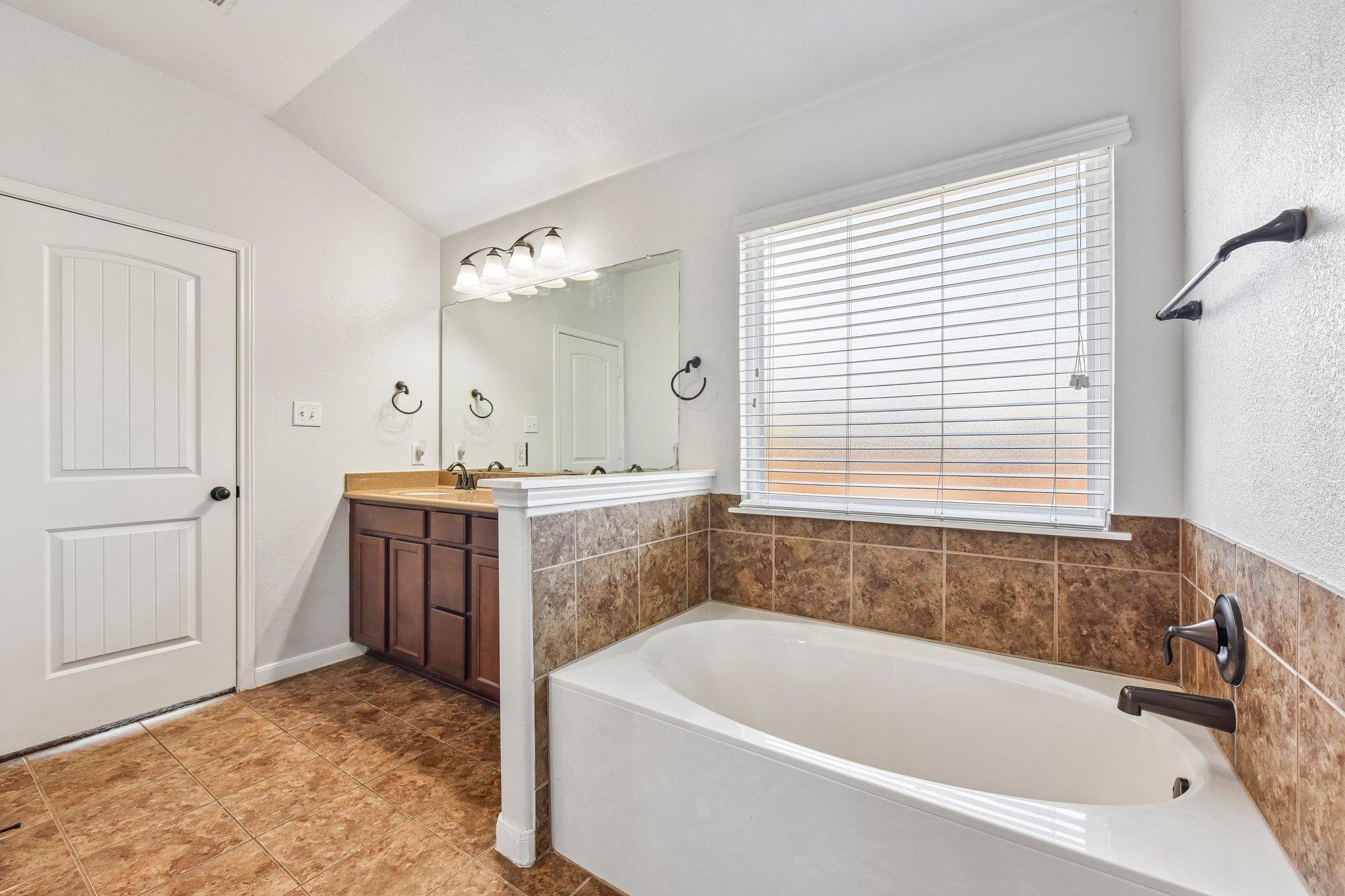 Bathroom with a white soaking tub, brown tile surround, and dark wood vanity beside a window.