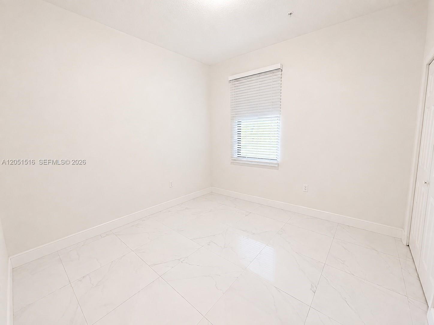 Vacant room with large white tile flooring and a window with blinds.