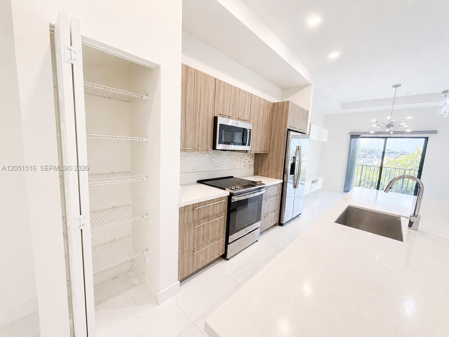 Modern kitchen with wood-grain cabinets, stainless appliances, and a large white island beside an open pantry.