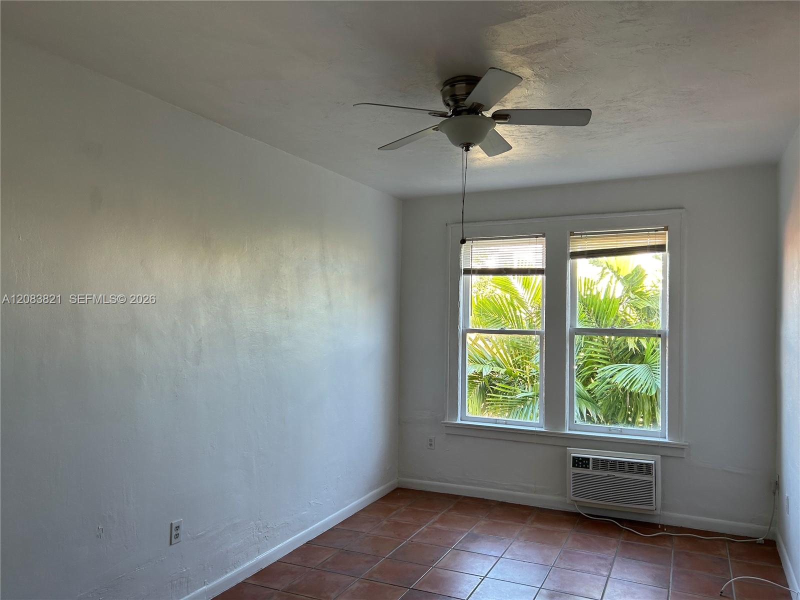 Empty room with terracotta tile floors, a ceiling fan, and two windows overlooking palms.