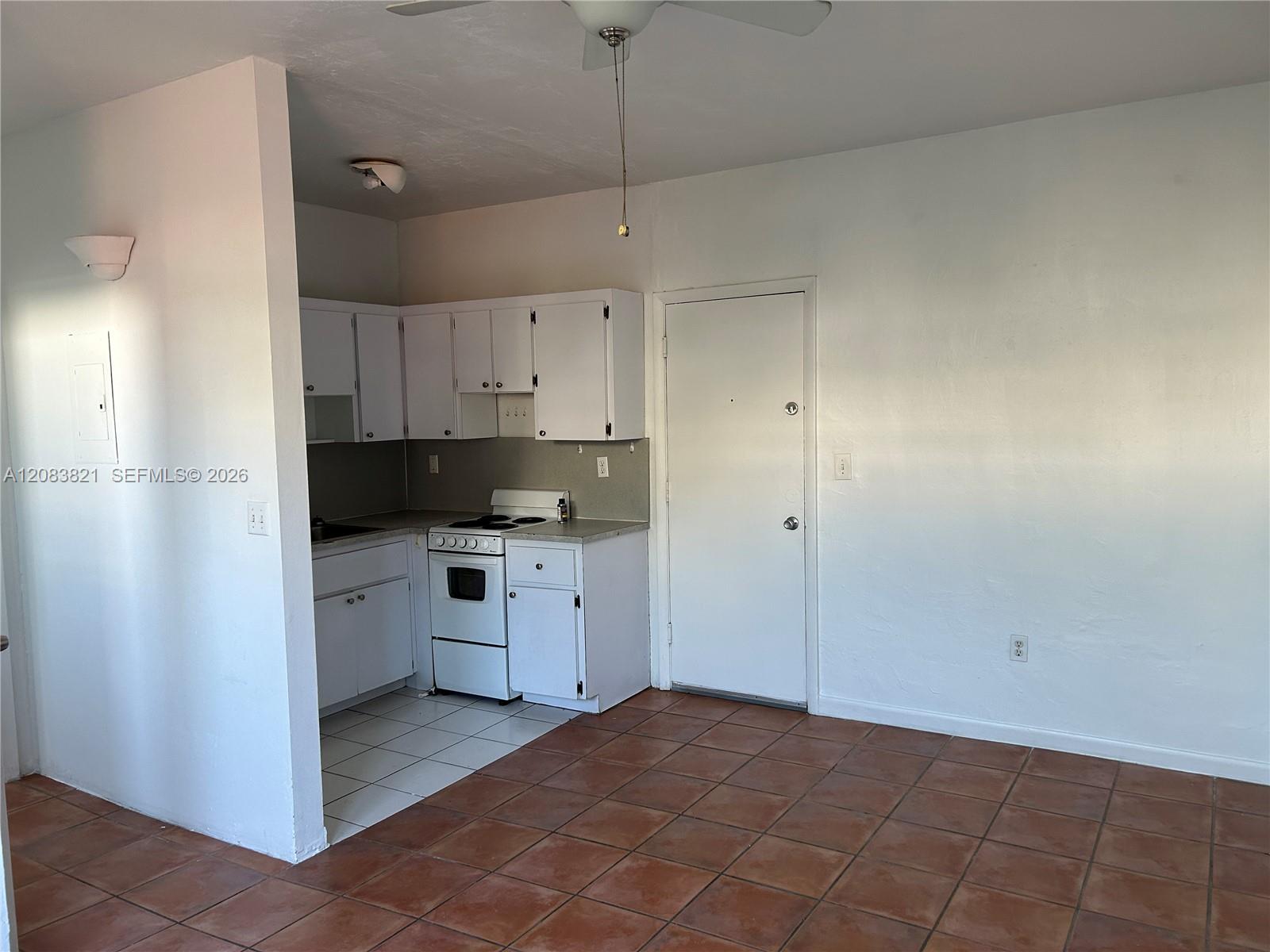 Small apartment kitchen with white cabinets and electric range, opening onto a tiled room with a closed door.