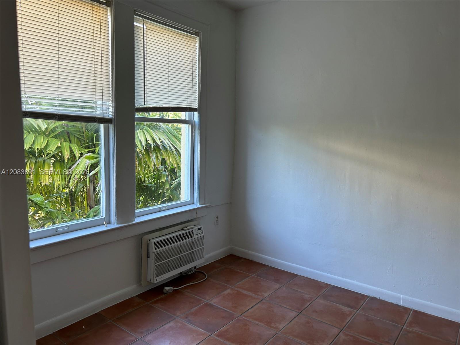 Empty tiled room with two shaded windows, a wall-mounted air conditioner, and tropical foliage outside.