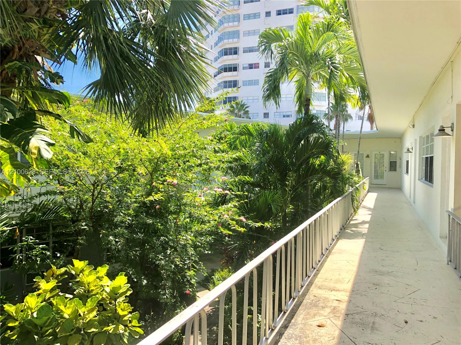 Exterior walkway beside a lush tropical courtyard with palm trees and a white residential building.