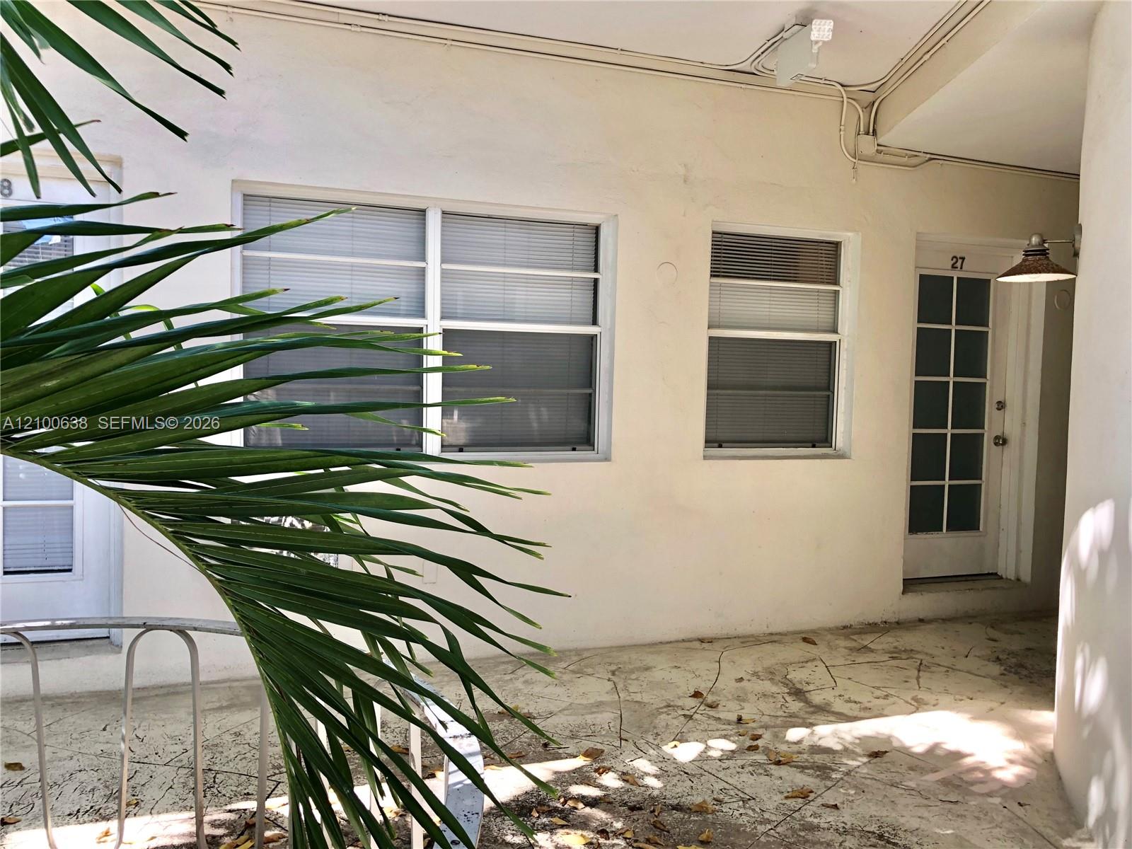 Unit 27 entrance along a tiled exterior walkway, partly screened by tropical palm fronds.