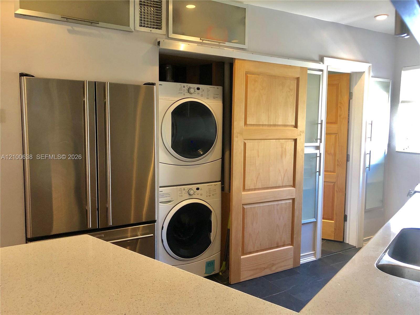 Kitchen laundry area with a stainless steel refrigerator and stacked washer and dryer behind a sliding wood door.