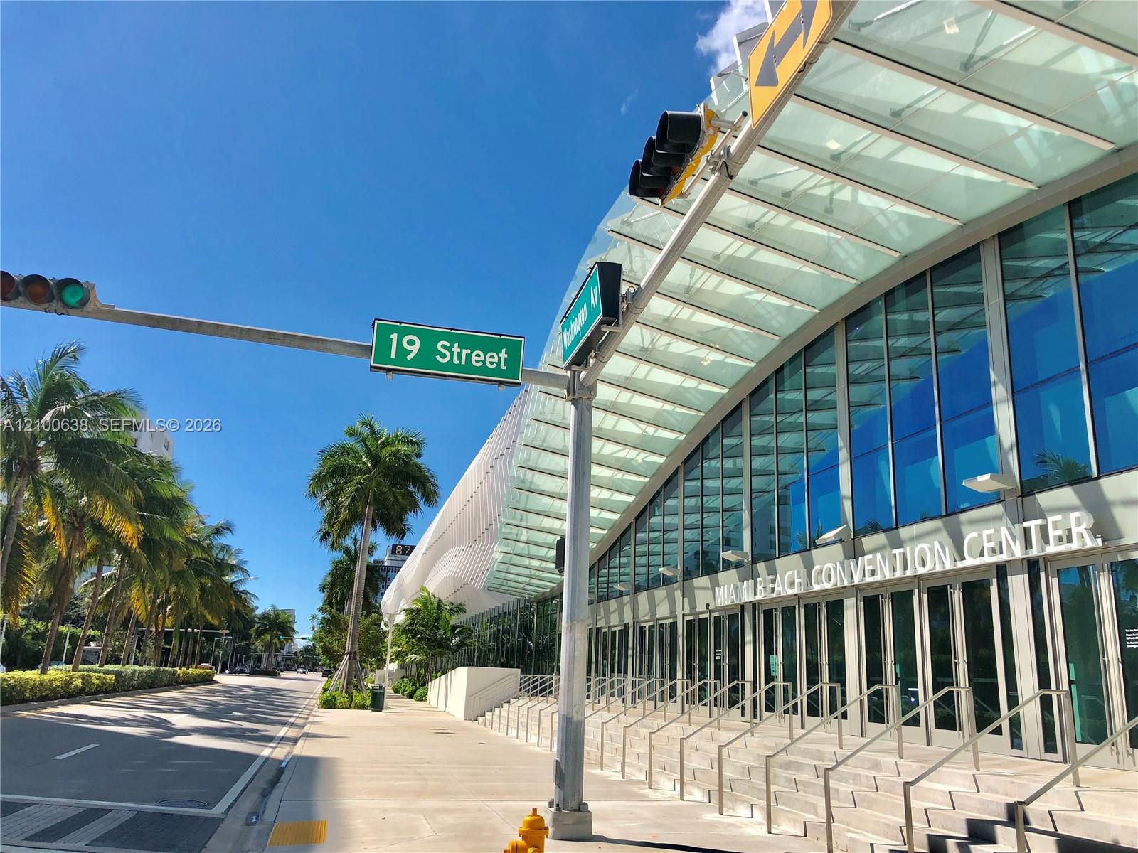 Miami Beach Convention Center entrance beside 19 Street and Washington Avenue, with glass facade and palm-lined sidewalk.