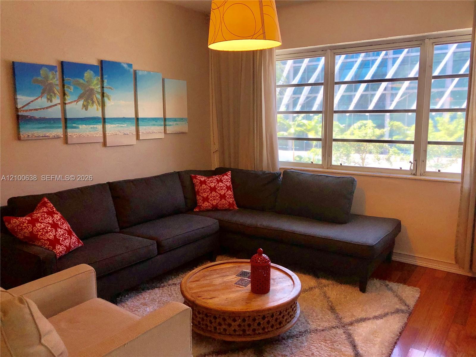 Living room with a dark sectional, beach-themed wall art, and large sunlit windows.