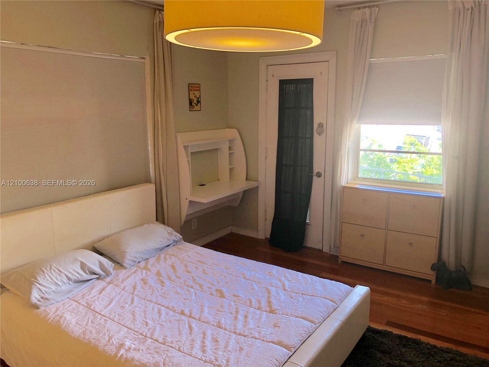 Bedroom with a white bed, hardwood floors, wall desk, and curtained window.