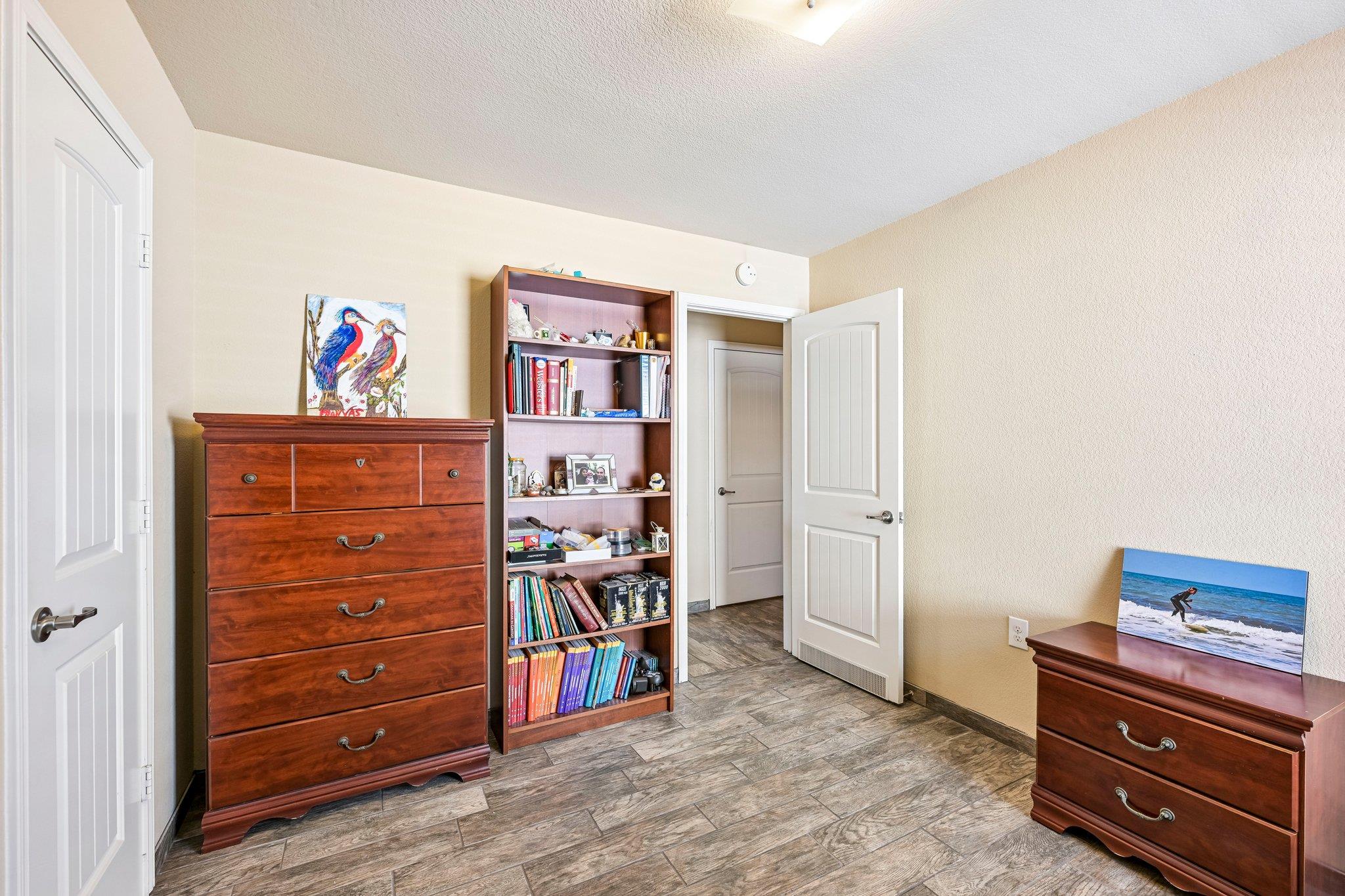 Bedroom with wood-look tile flooring, wooden dressers, and a tall bookshelf beside an open doorway.