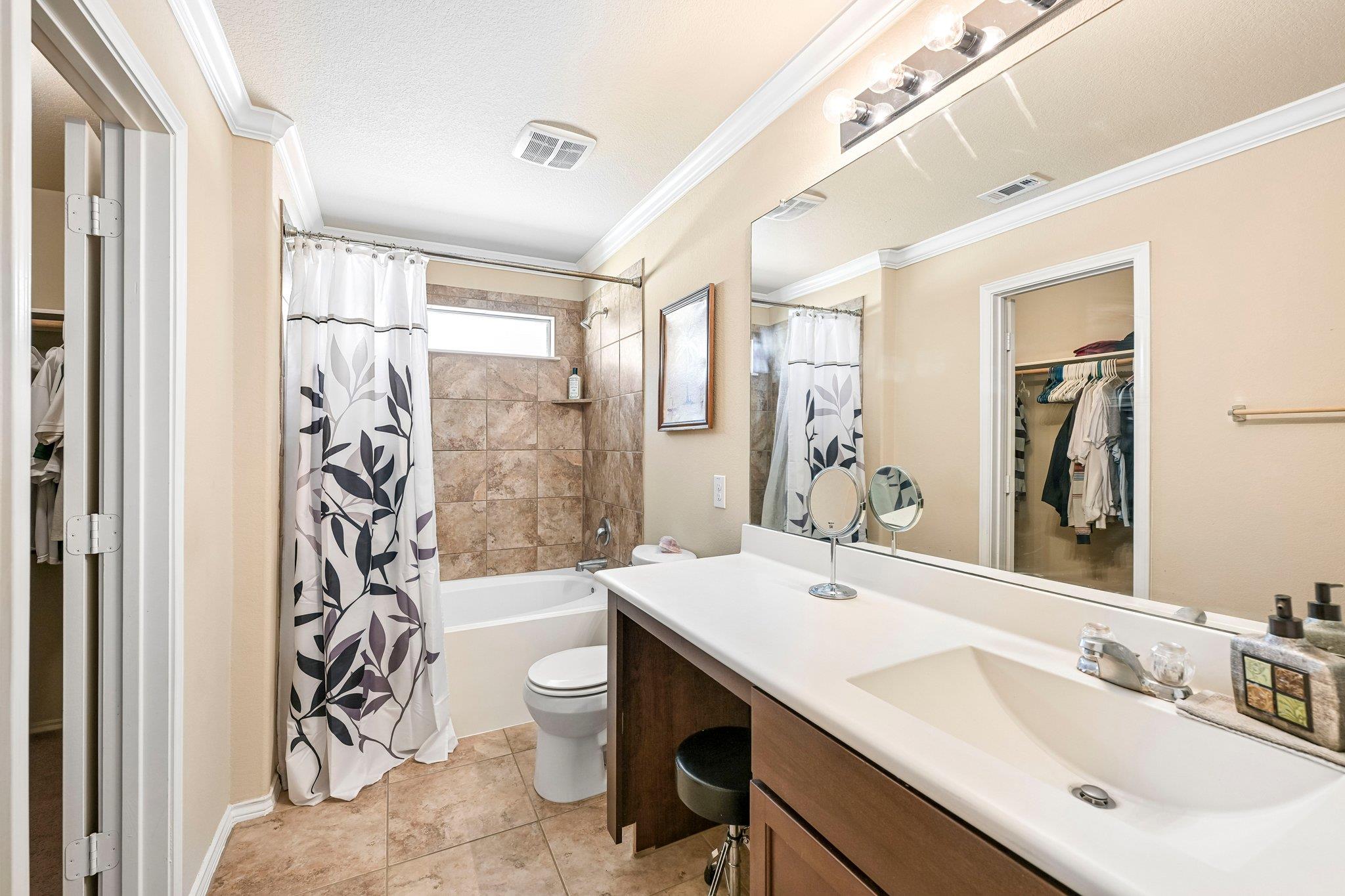 Bathroom with a tiled tub and patterned shower curtain beside a long double-sink vanity and mirrored wall.
