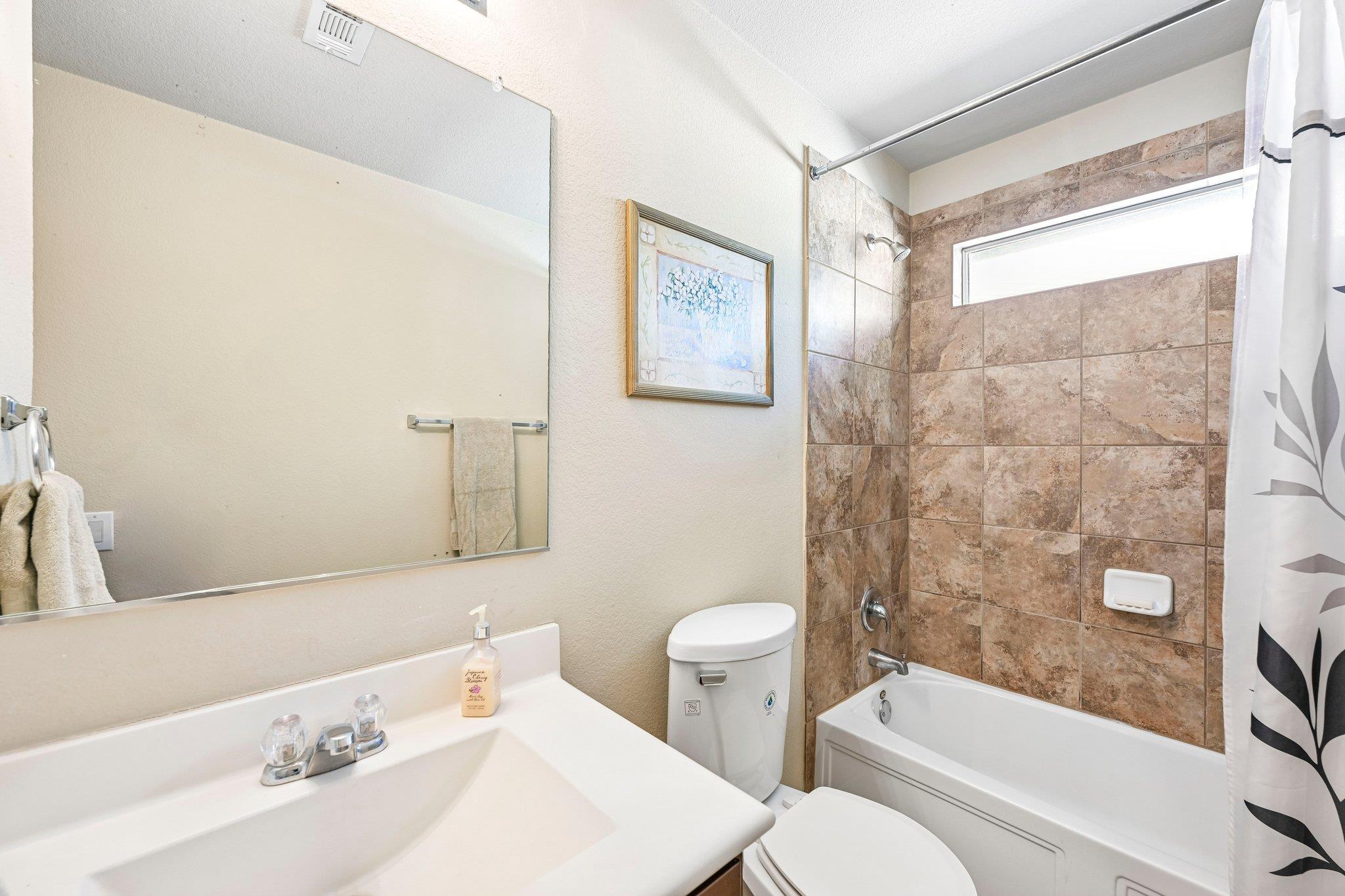 Bathroom with a white vanity and a bathtub surrounded by brown tile beneath a high window.