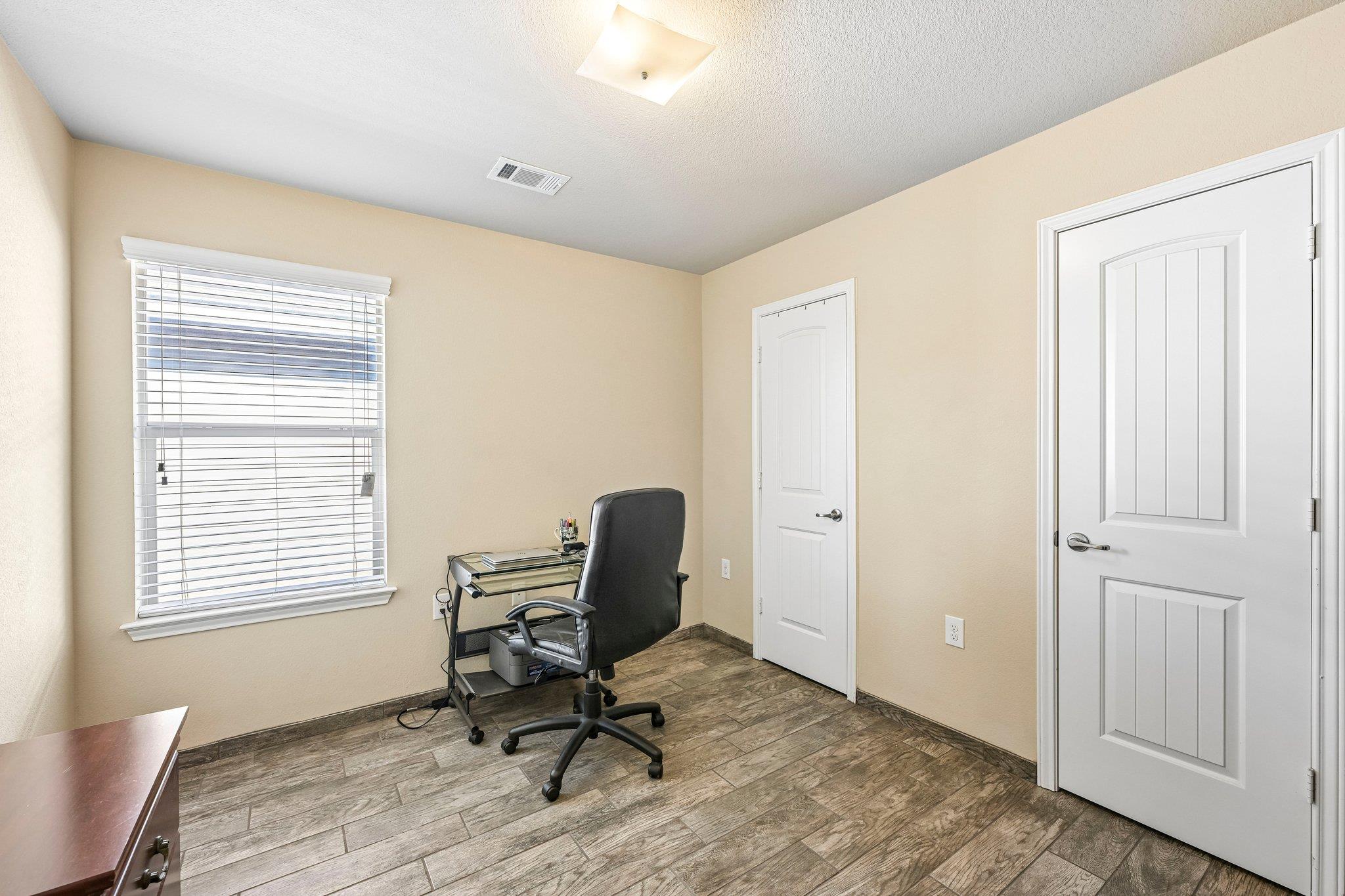 Home office with a glass desk, black chair, wood-look tile floor, and window with blinds.