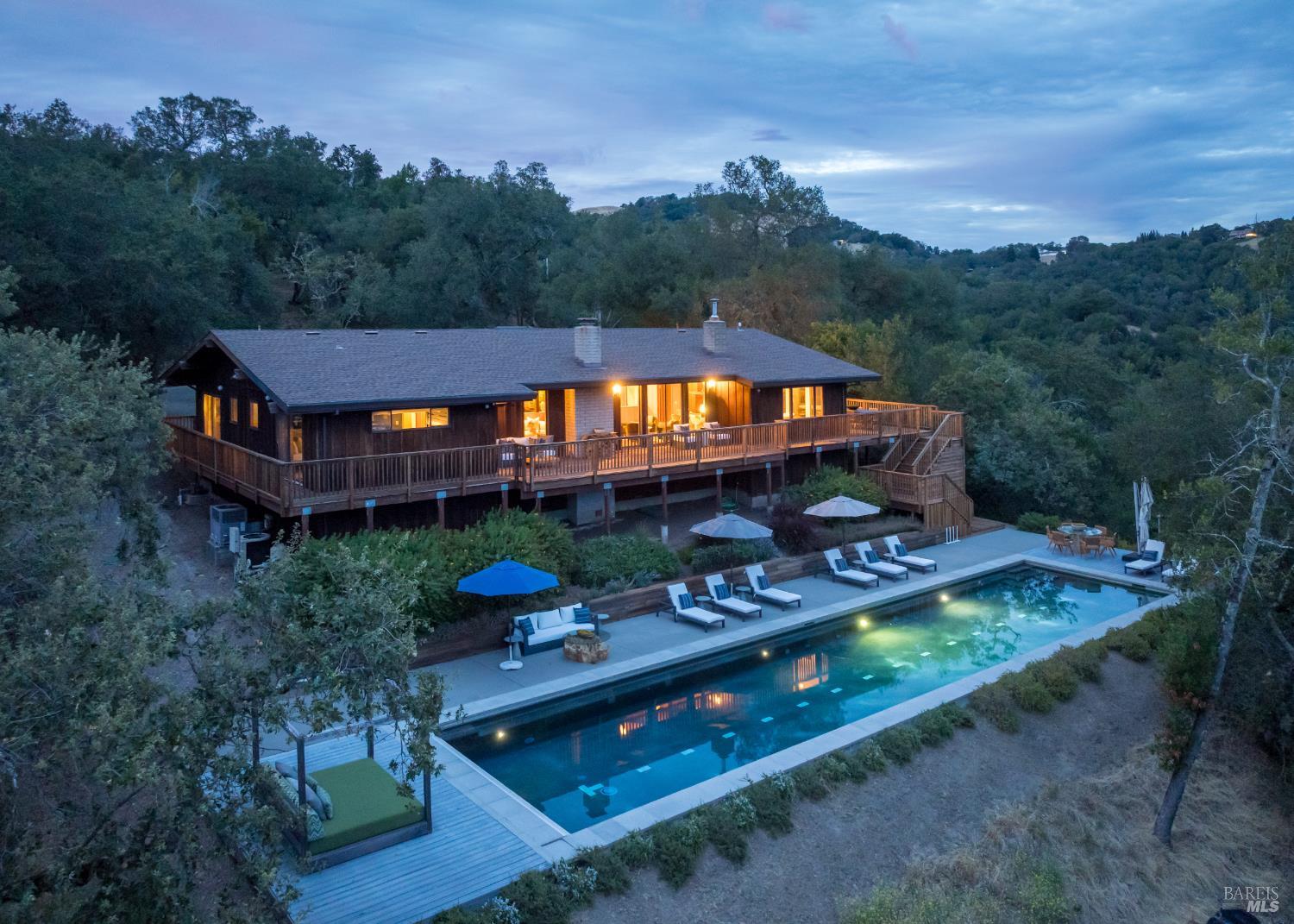 Wooded hillside home with a wraparound deck and illuminated swimming pool at dusk.