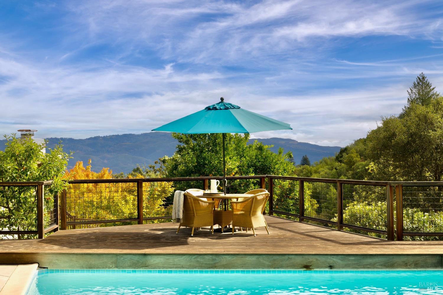 Swimming pool and dining deck with a teal umbrella overlooking wooded hills and distant mountains.