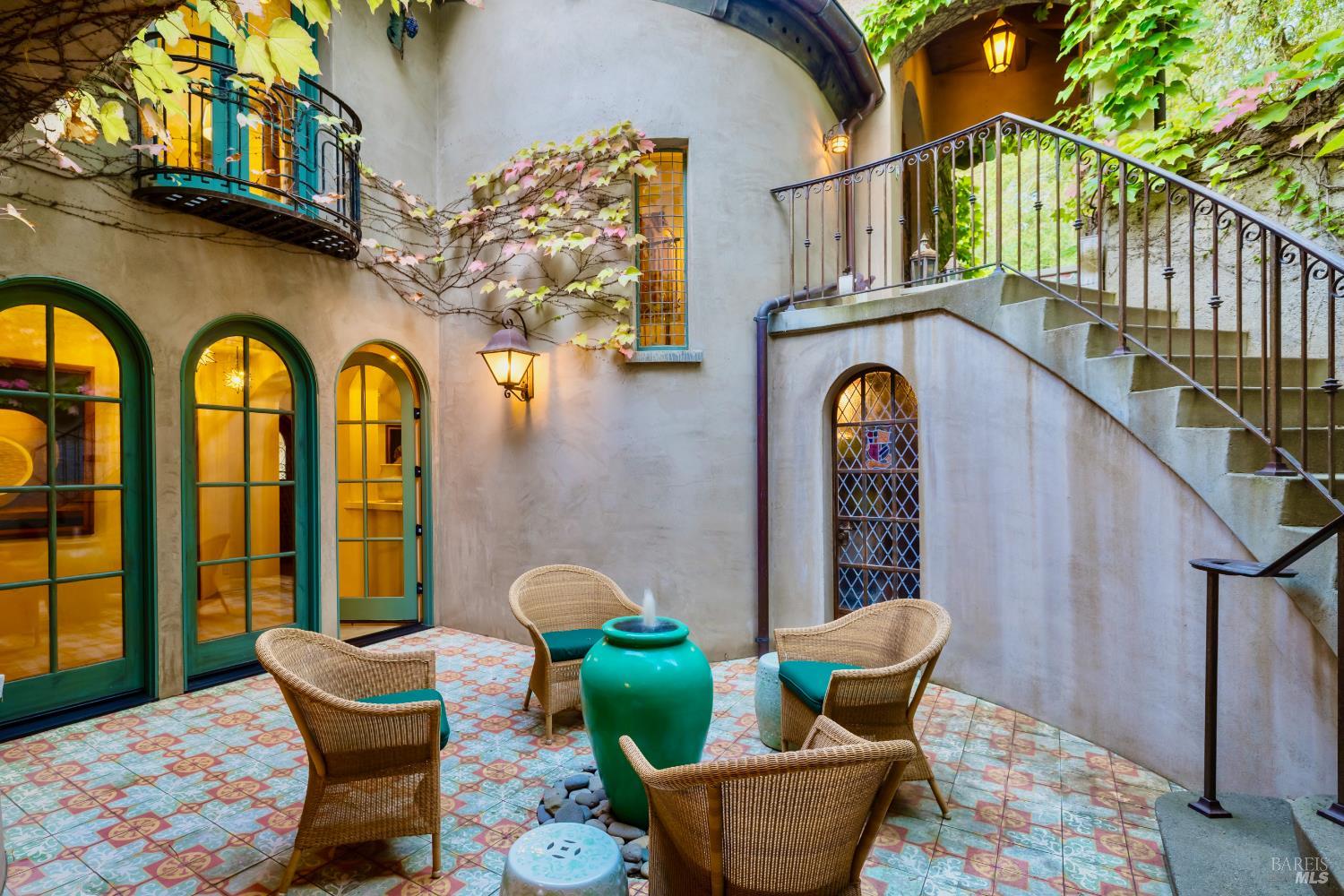 Courtyard with patterned tile, green arched doors, wicker seating, climbing vines, and a curved staircase.