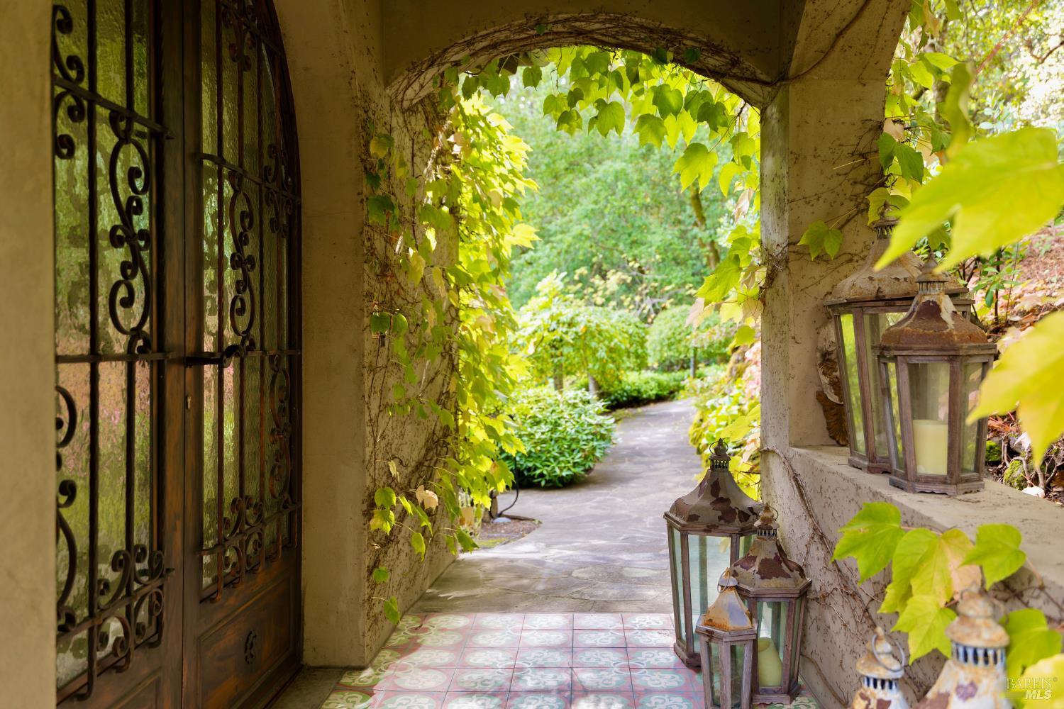 Vine-covered entryway with an ornate iron door, patterned tile, and lanterns along a garden walkway.