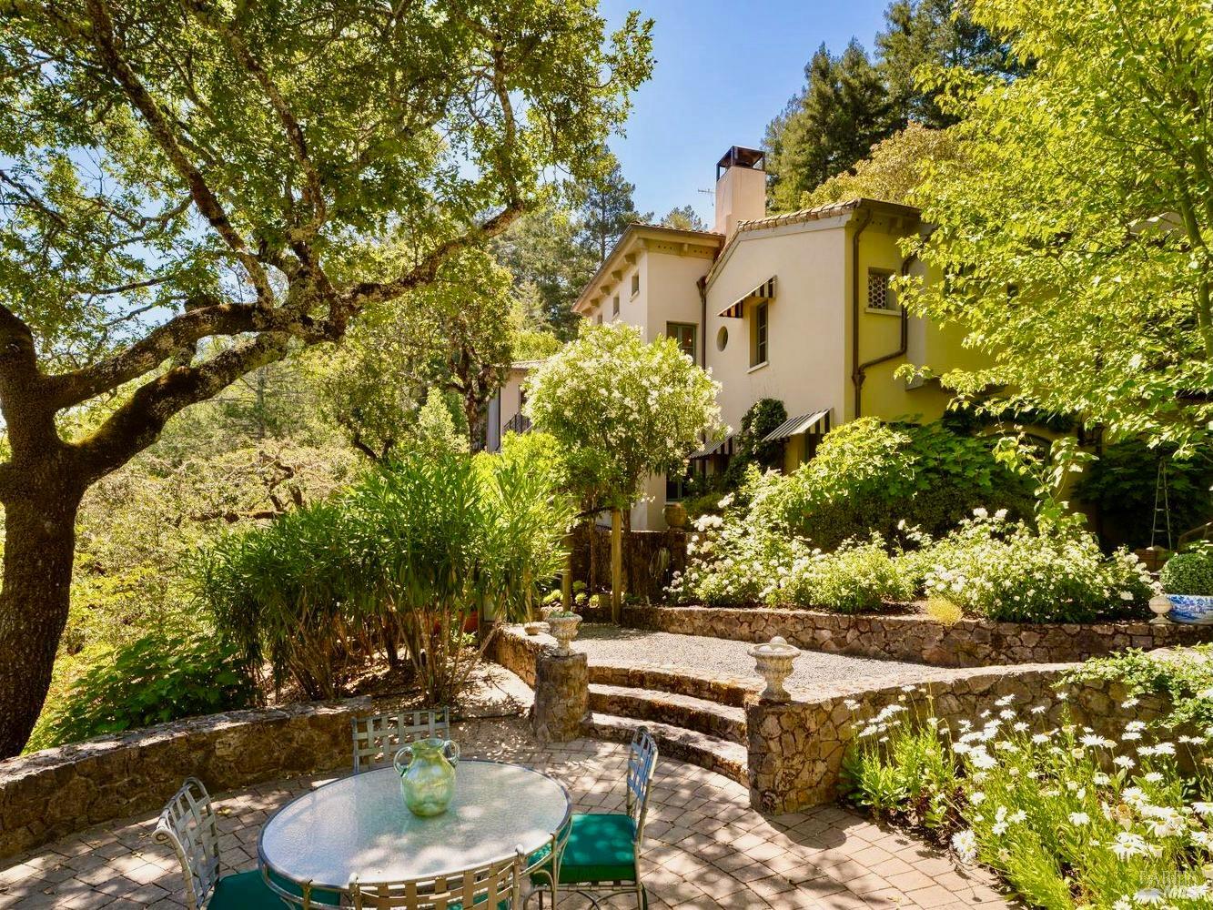 Stucco home behind a landscaped stone terrace with outdoor dining furniture and flowering plants.
