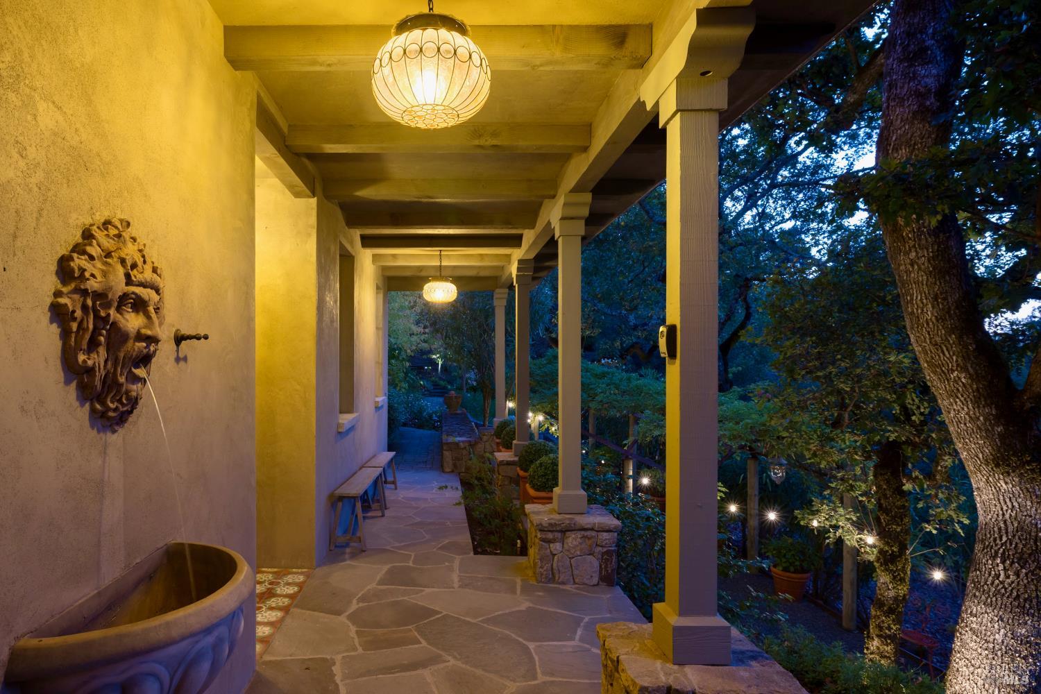 Covered stone porch with glowing pendant lights, a sculpted wall fountain, and a wooded garden at dusk.