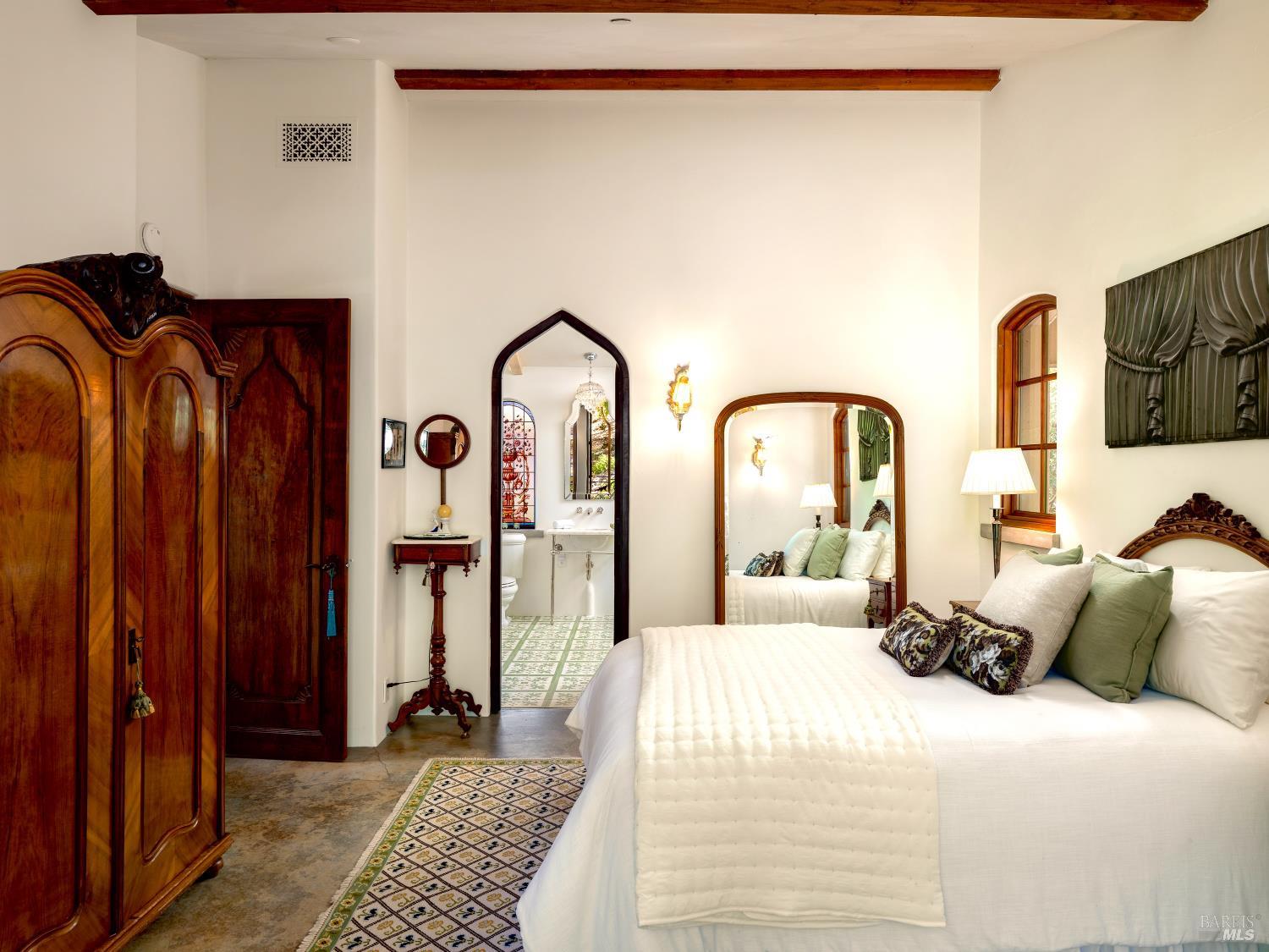 Bedroom with a white bed, carved wooden furniture, exposed beams, and an arched ensuite doorway.