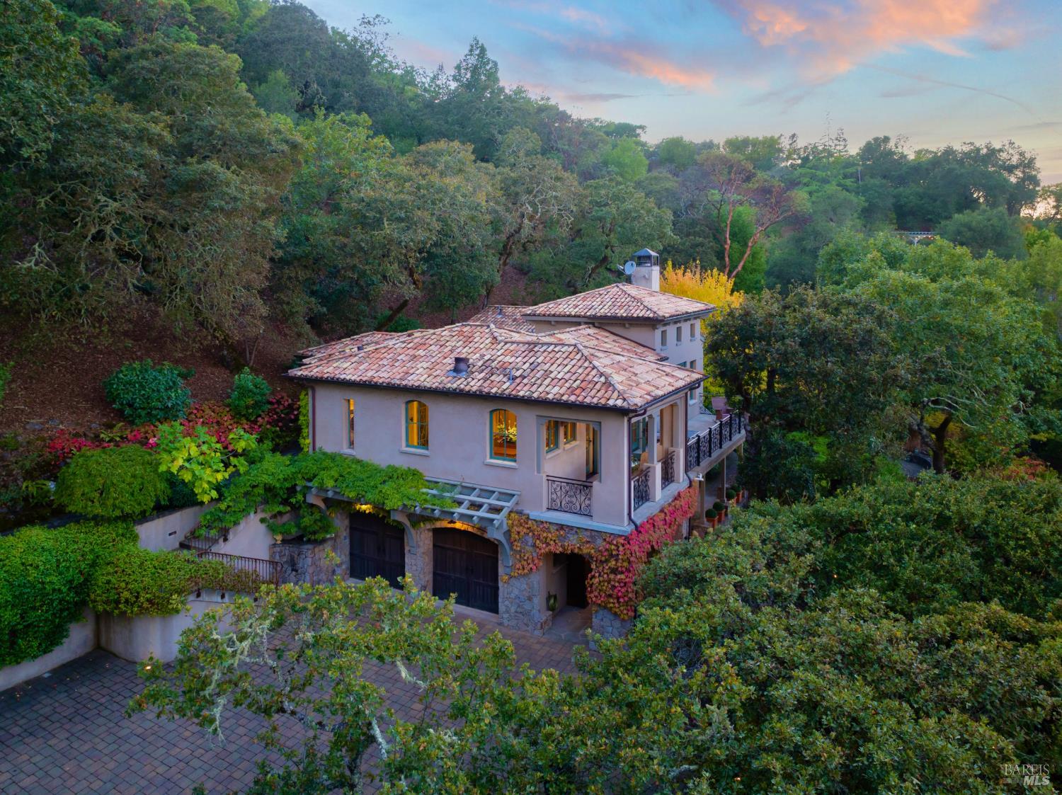 A tile-roofed home with balconies and stone garage frontage nestled among trees at dusk.