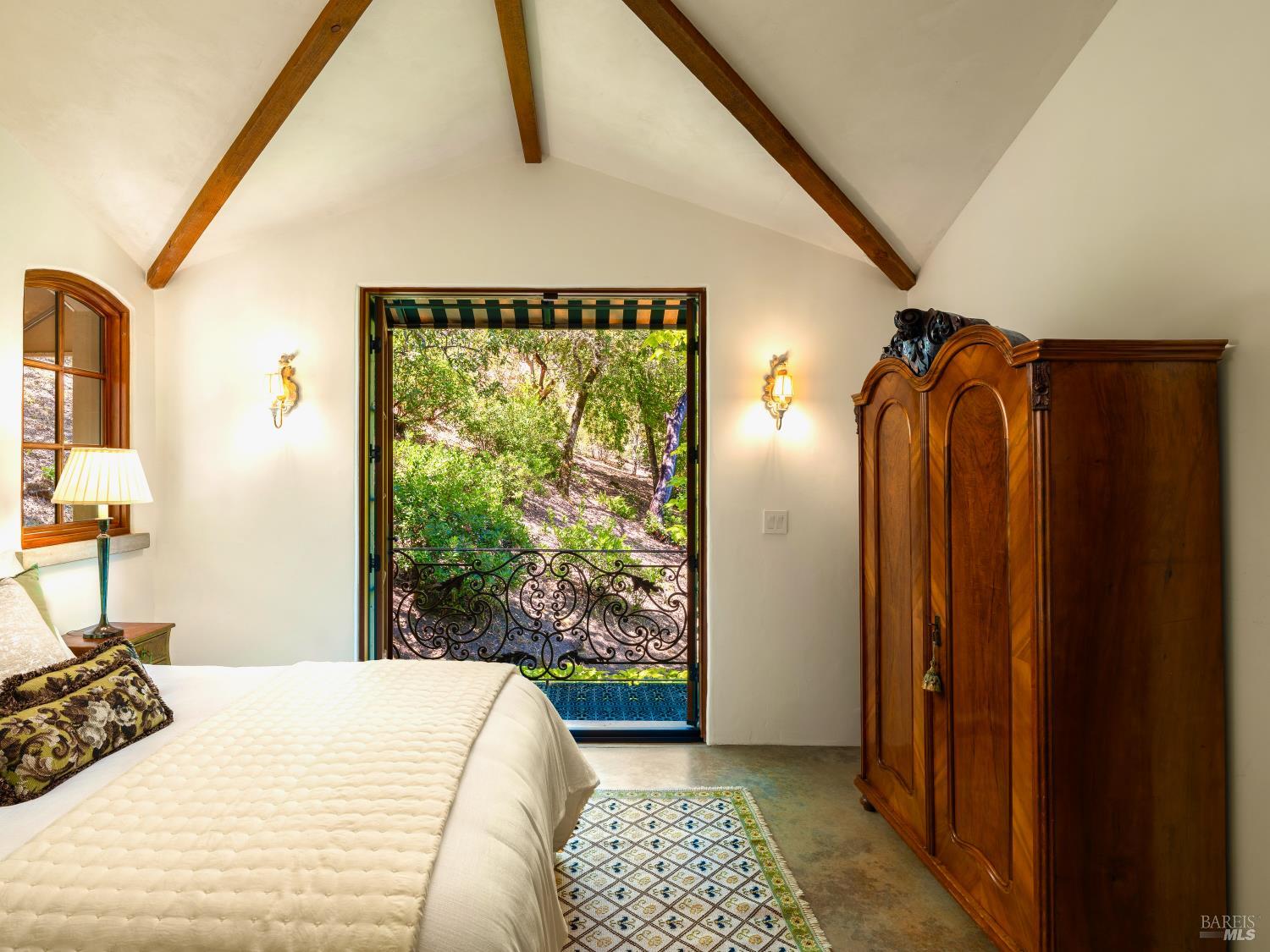 Bedroom with exposed wooden beams, a carved wardrobe, and a doorway overlooking a leafy garden.