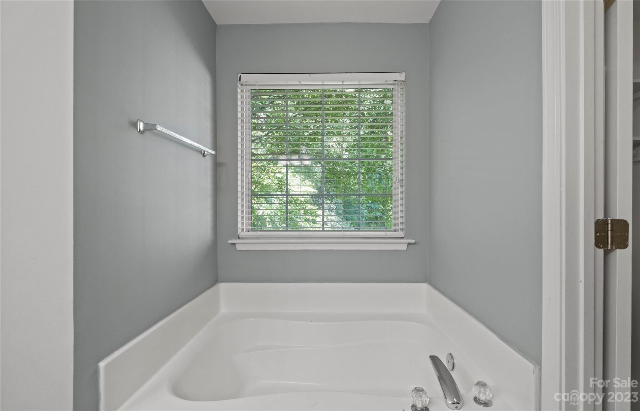 Built-in bathtub beneath a window with white blinds in a gray bathroom.