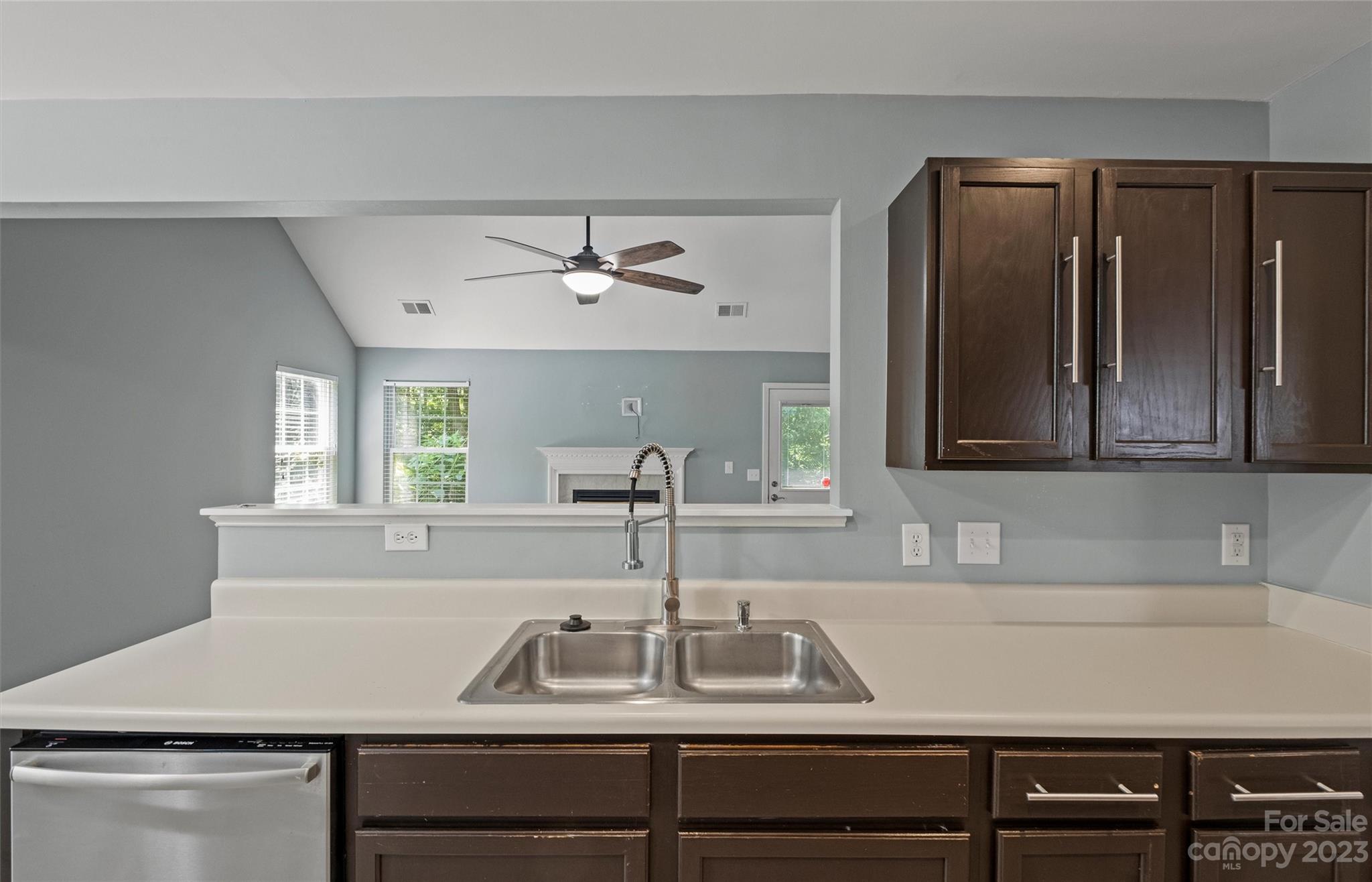 Kitchen sink and white countertop with dark cabinets overlooking a living area and fireplace.